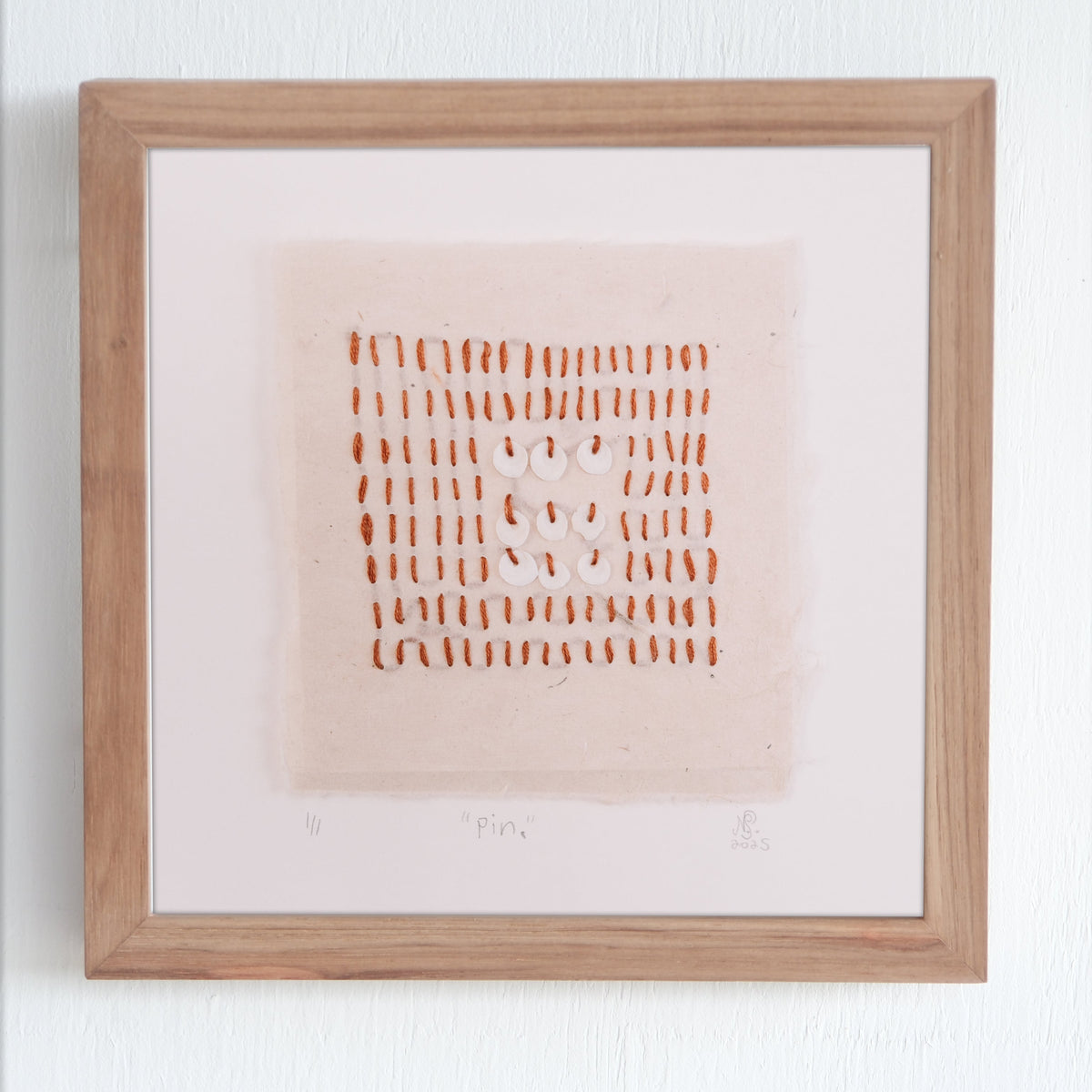 A framed abstract minimal art work featuring a grid of small terracotta coloured naturally dyed vertical stitches with a central cluster of white circular shells, on lokta Nepalese paper within a natural wood frame against a white wall.
