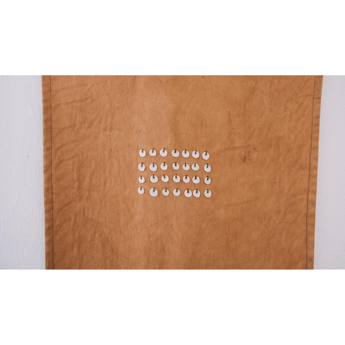 A close up square hand dyed brown textured fabric  panel hanging against a white wall, with a small grid of white oval shells arranged in three rows at the center.